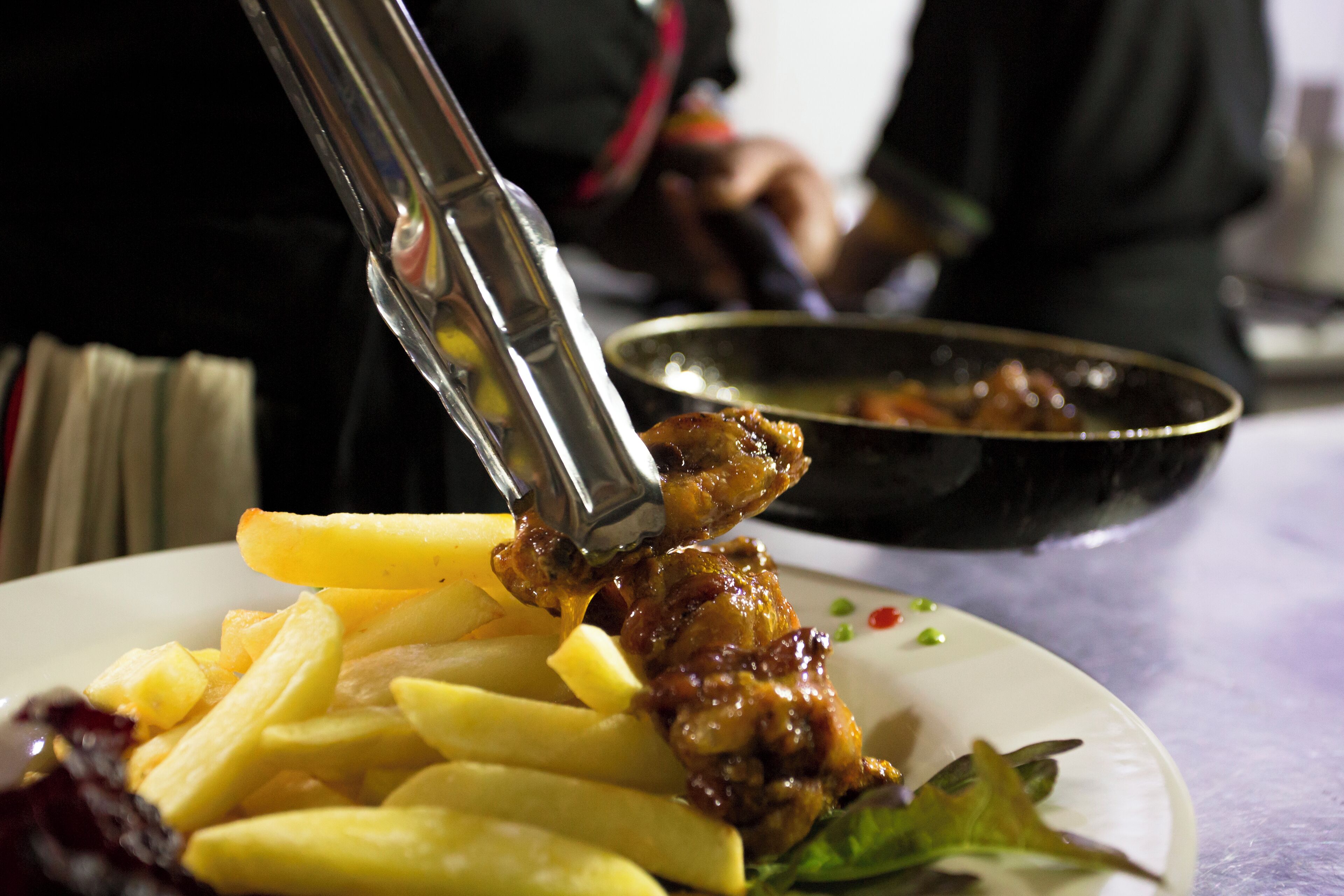 Une assiette avec des frites et un plat de viande, un bol à l’arrière-plan et une personne à l’arrière-plan.