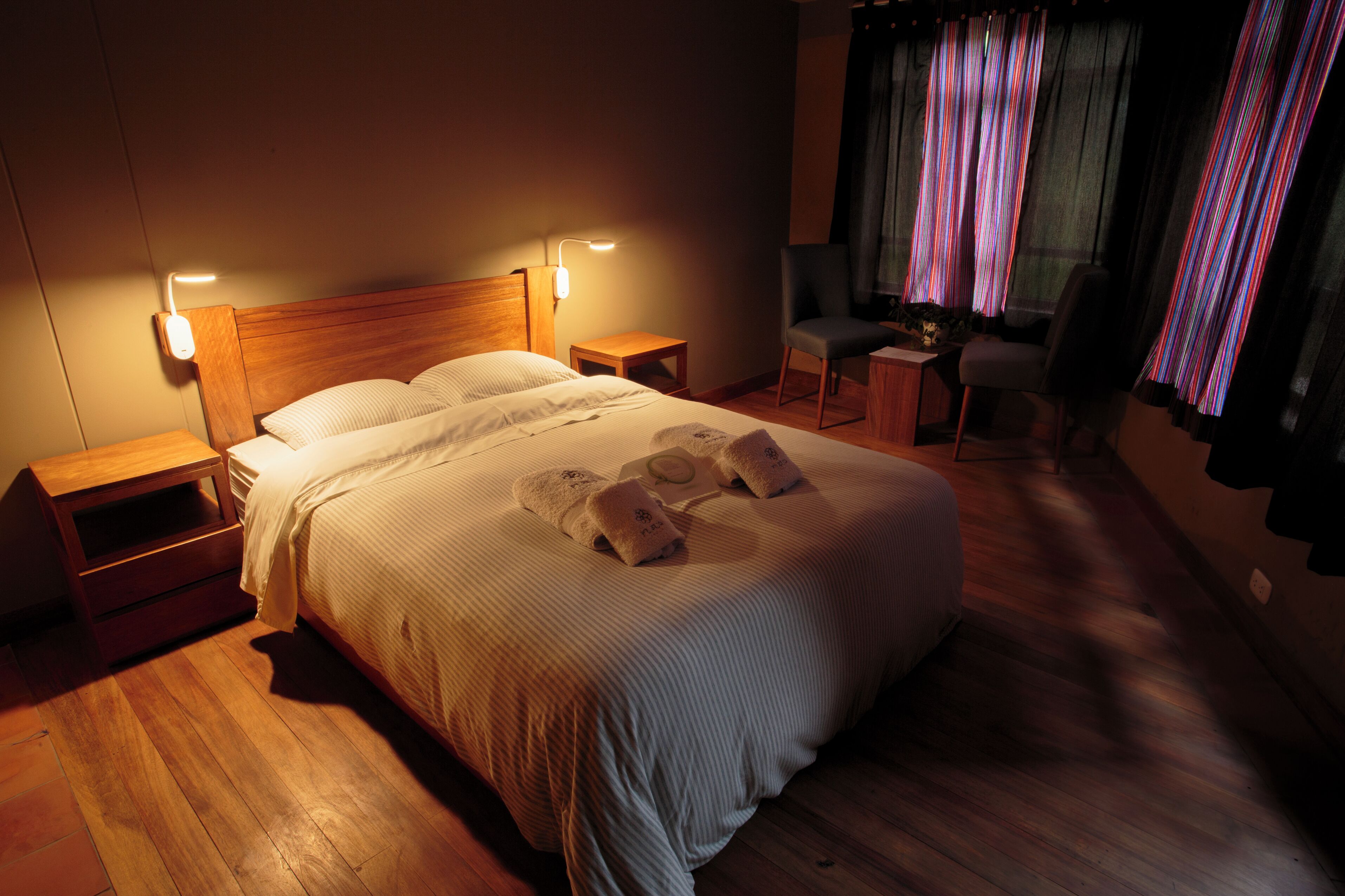 A neatly made bed with pillows and a blanket, wooden nightstands, a small desk with a chair, and a window with curtains.