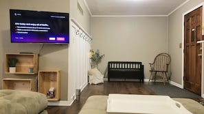A living room with a flat-screen TV displaying a Netflix sign, a wooden bench, a cushioned chair, a small plant, and a wooden shelf with a tissue box.