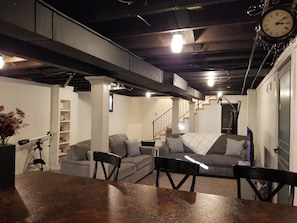 A room with a wooden table, two grey sofas, a black chair, a white pillar, a staircase, and a clock on the wall.