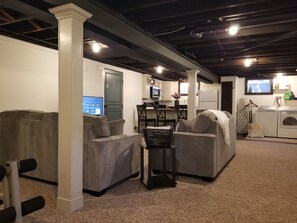 A room with two sofas, a small table, a washing machine, and a television displaying a streaming service.