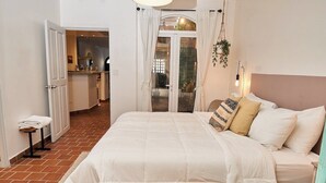 Ein Schlafzimmer mit einem großen Bett, einem kleinen Nachttisch und Blick auf eine Küche und einen Essbereich durch eine offene Tür.