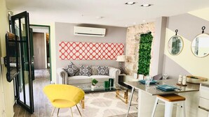 Ein modernes Wohnzimmer mit einer Couch, einem gelben Sessel, einem Esstisch mit Hockern und einer Küchenzeile mit grüner Wand und einem gemusterten Teppich.