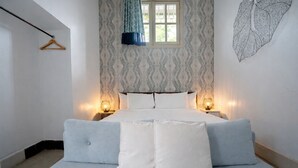 Un dormitorio con cama, dos mesitas de noche con lámparas, una ventana con cortinas y una pared con estampado de hojas.