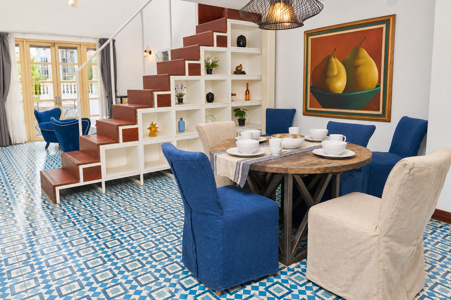 Un coin repas avec une table ronde dressée pour quatre personnes, entourée de chaises bleues et beiges. Un escalier avec des marches en bois et des étagères blanches est visible à l’arrière-plan.