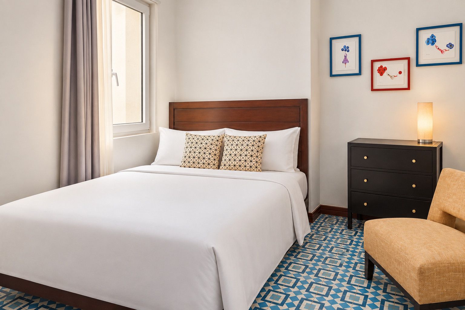 Camera d'albergo con un letto grande, una cassettiera, una sedia e un comodino con una lampada.
