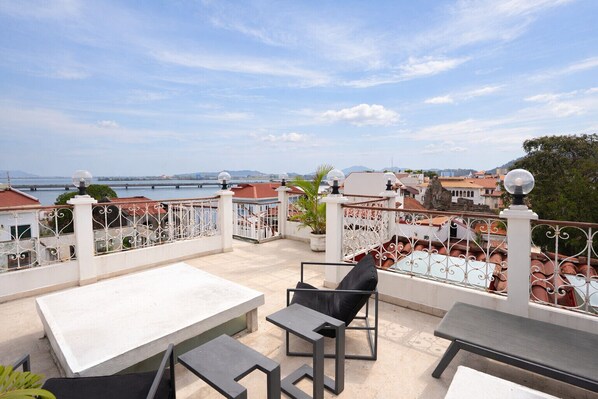 A rooftop terrace with outdoor seating, a view of the sea, and a bridge in the distance.