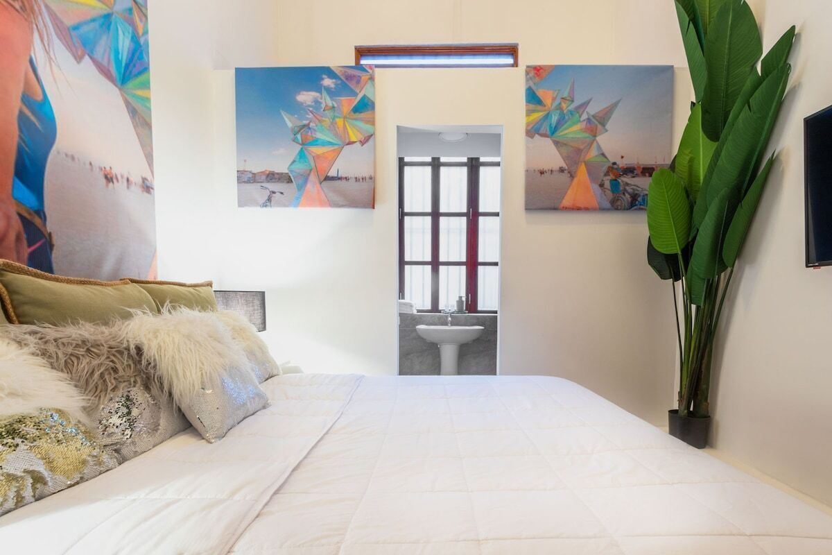 A bedroom with a bed, pillows, a large plant, and three paintings on the wall.