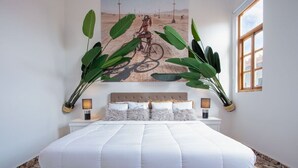 Una cama bien hecha con edredón blanco, cabecera con panel de madera y una obra de arte en la pared que muestra una bicicleta.