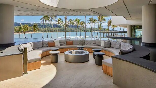Un área de descanso moderna con disposición circular de asientos, una chimenea y vistas a la playa y palmeras.