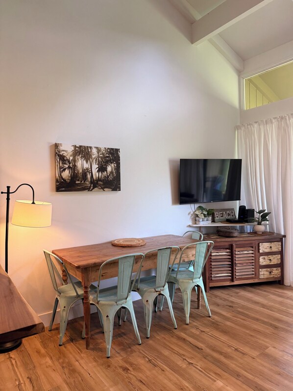 A dining area with a wooden table and four chairs, a painting of palm trees, a lamp, a television, and a shelf with a 'BEACH' sign.