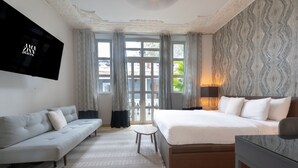 Habitación de hotel moderna con una cama grande, un sofá, un televisor montado en la pared y un balcón con cortinas.