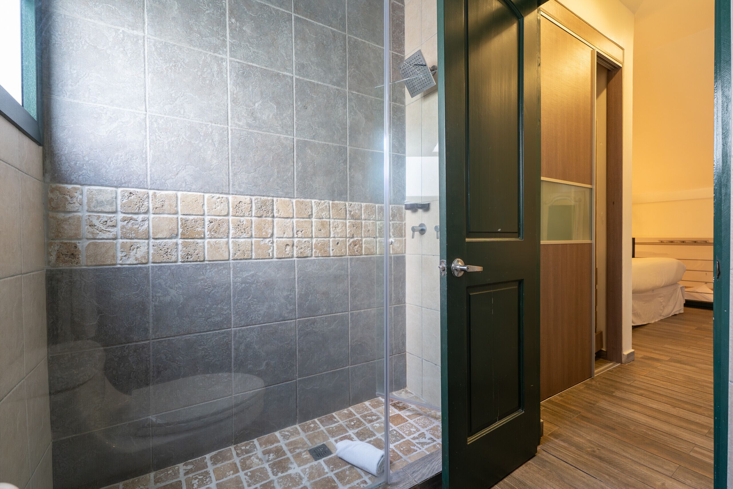 Una ducha con puerta de vidrio, pared con azulejos y puerta de madera que da a un dormitorio.