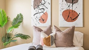 A neatly made bed with a white comforter and two rolled towels, accompanied by a large green plant and two abstract paintings on the wall.