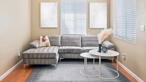 A living room with a grey sectional sofa, a round coffee table, and a grey rug. There are three framed artworks on the wall and a window with blinds.