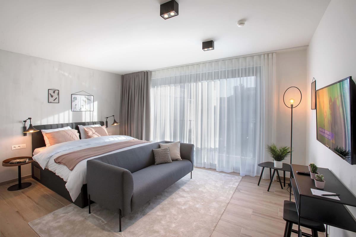 Ein modernes Schlafzimmer mit Bett, Nachttischen, einem Schreibtisch, einem Sessel und einem großen Fenster mit transparenten Vorhängen.