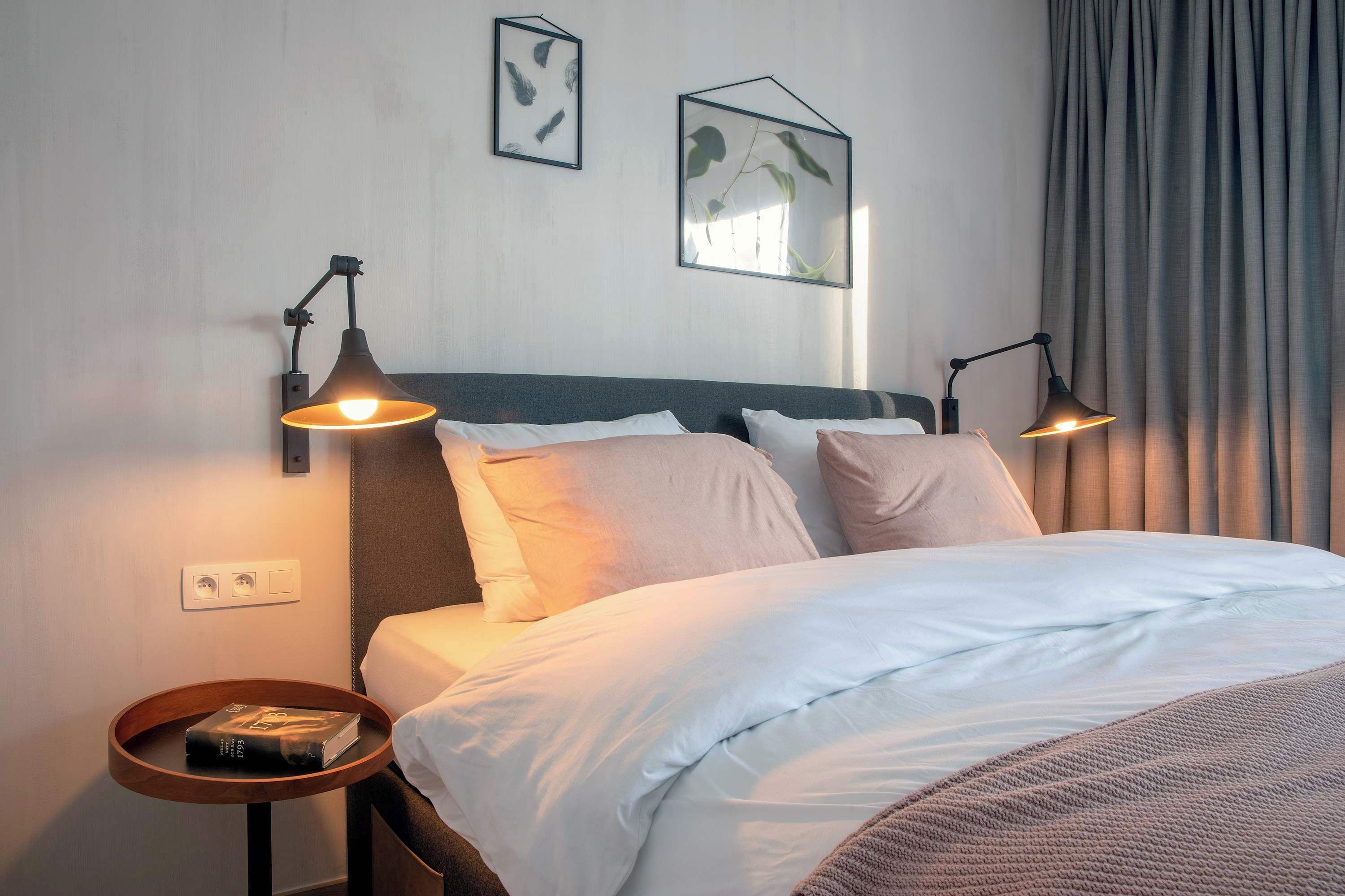 Een keurig opgemaakt bed met witte lakens, twee bedlampjes, een klein houten nachtkastje met een boek, en een lijst met een plant aan de muur.