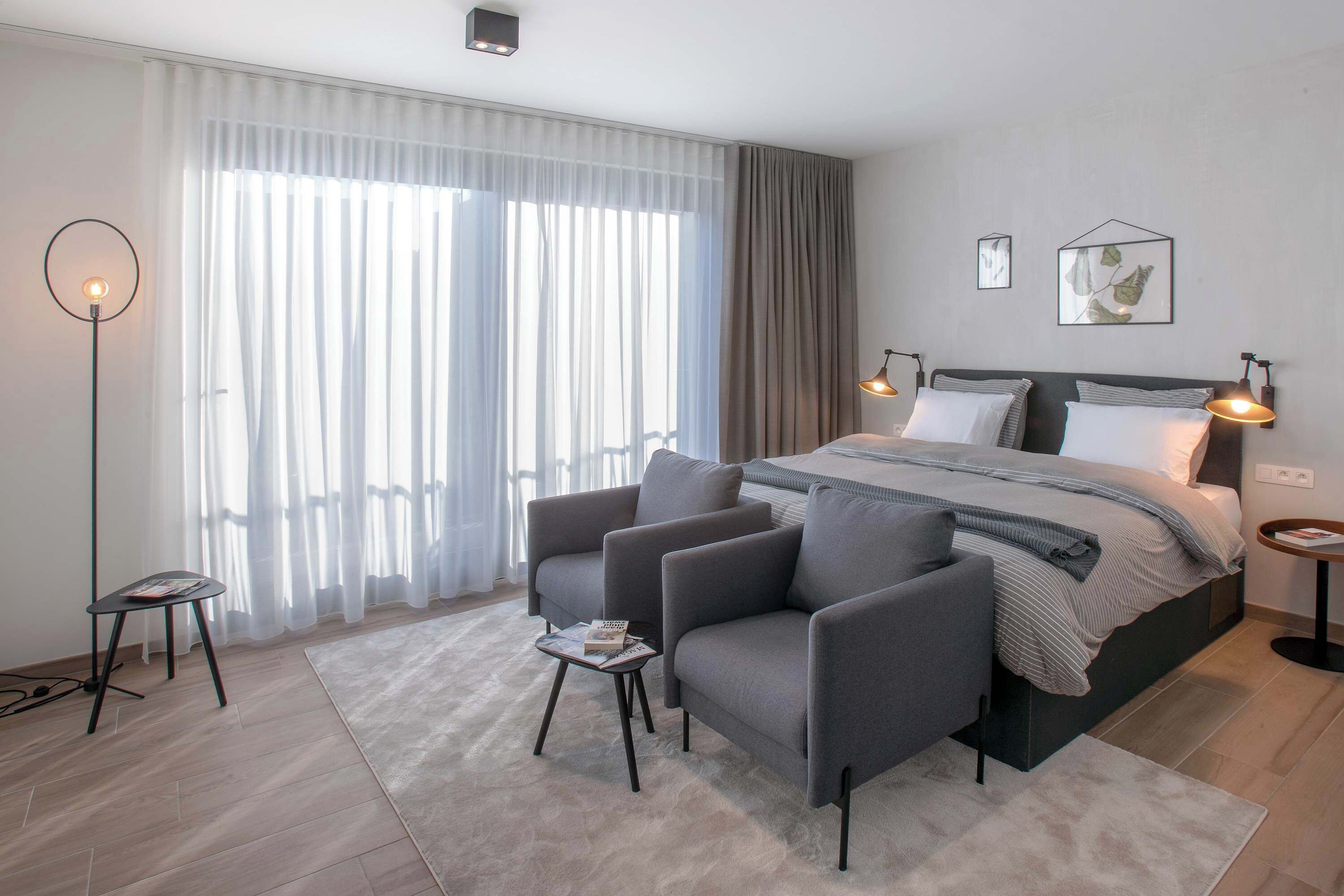 Una habitación de hotel moderna con una cama grande, sillones grises, una mesita y una lámpara de pie.