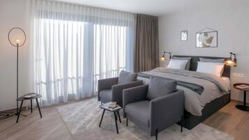 Una habitación de hotel moderna con una cama grande, sillones grises, una mesita y una lámpara de pie.