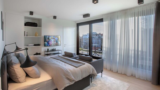 A modern bedroom with a large bed, a TV, a bookshelf, and a view of buildings through sheer curtains. - Q-Stay (Antwerp)