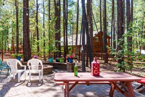 En stuga av trä, omgiven av höga träd, med ett picknickbord och stolar i förgrunden.