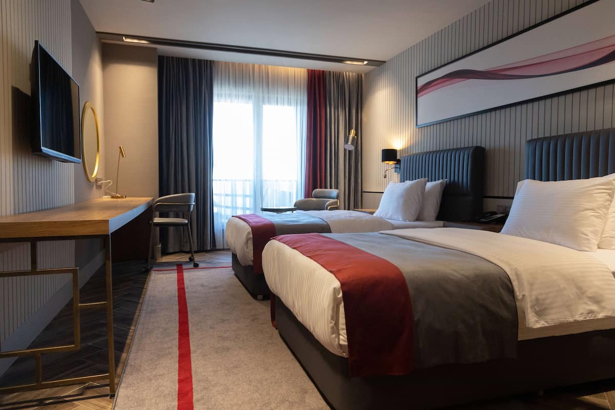 Une chambre d’hôtel avec deux lits, un bureau, une chaise, une télévision et une grande fenêtre avec des rideaux.