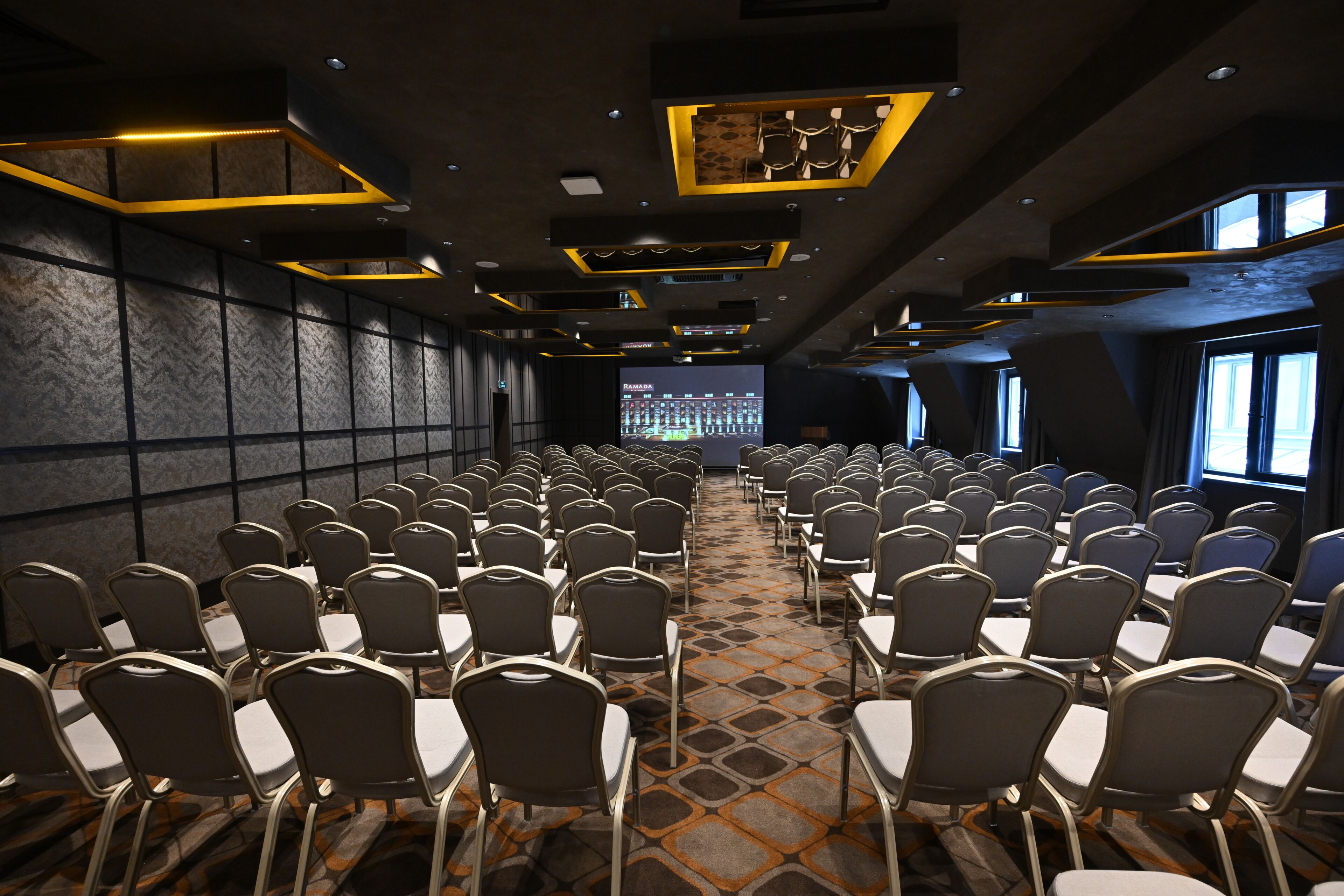 A conference room with rows of chairs, a patterned carpet, and a large screen at the front.
