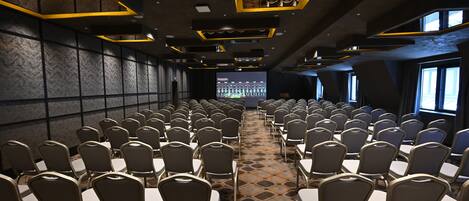 A conference room with rows of chairs, a patterned carpet, and a large screen at the front.