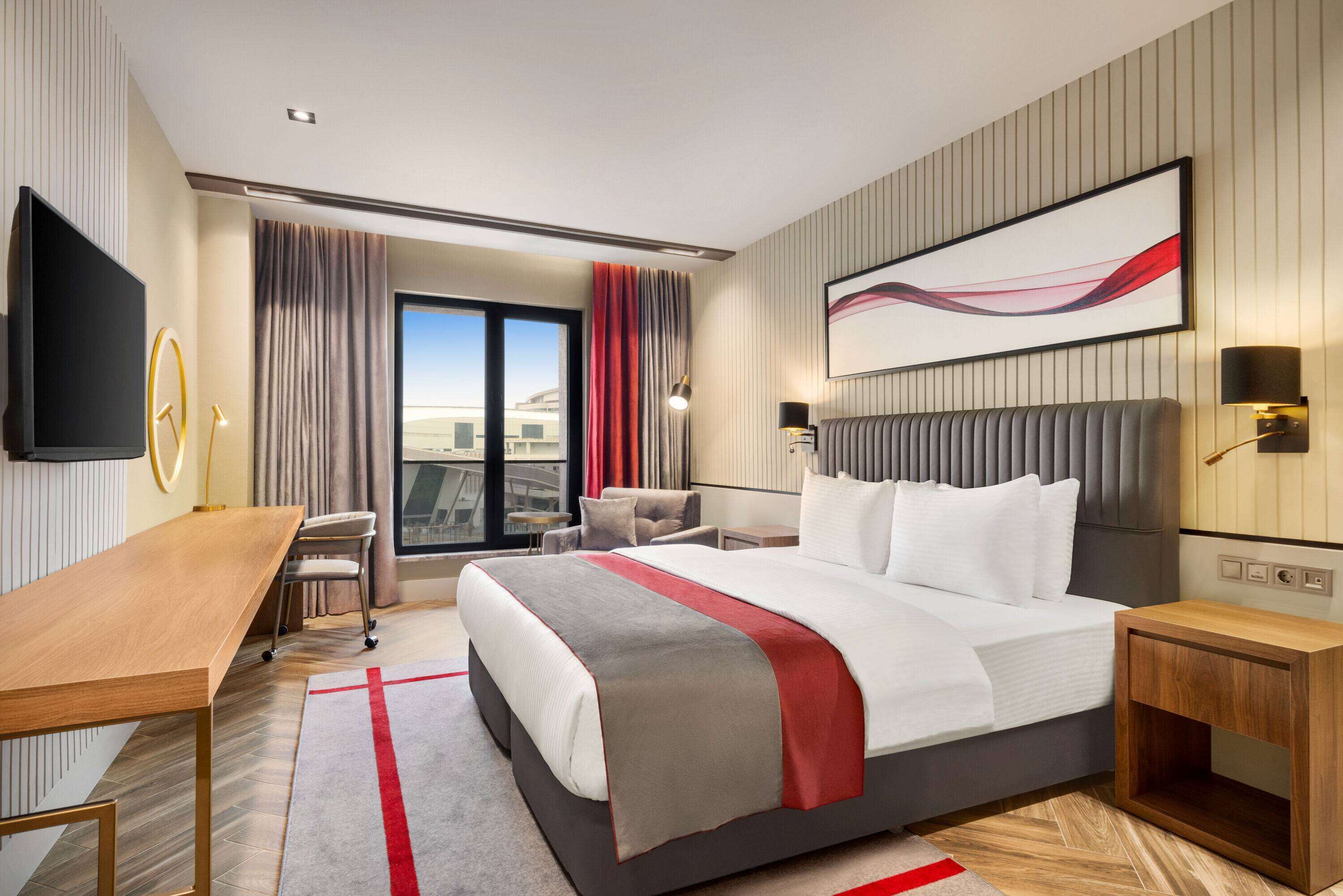 A modern hotel room with a large bed, a desk, a chair, and a view of the city.