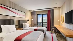 Ein Hotelzimmer mit zwei Betten, einem Schreibtisch, einem Fernseher und Blick auf die Stadt.