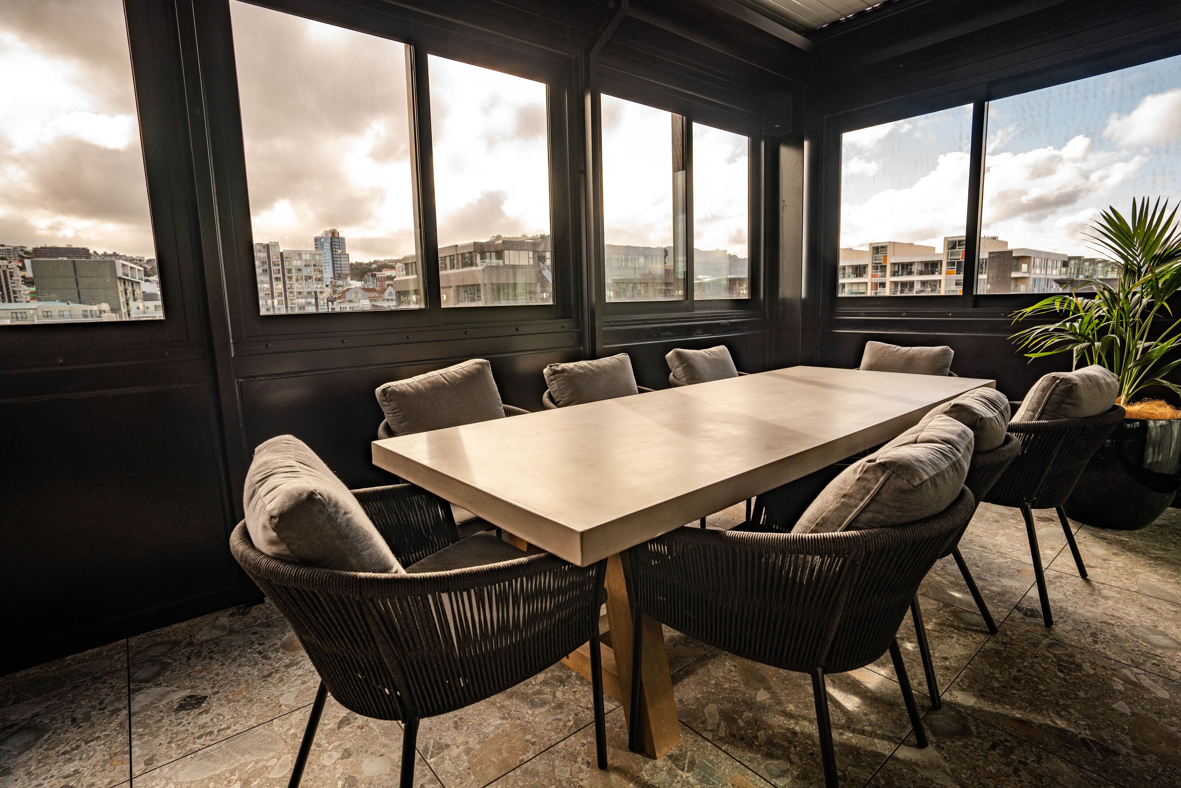 A sunlit room with a large table and chairs, large windows with a city view, and a potted plant.