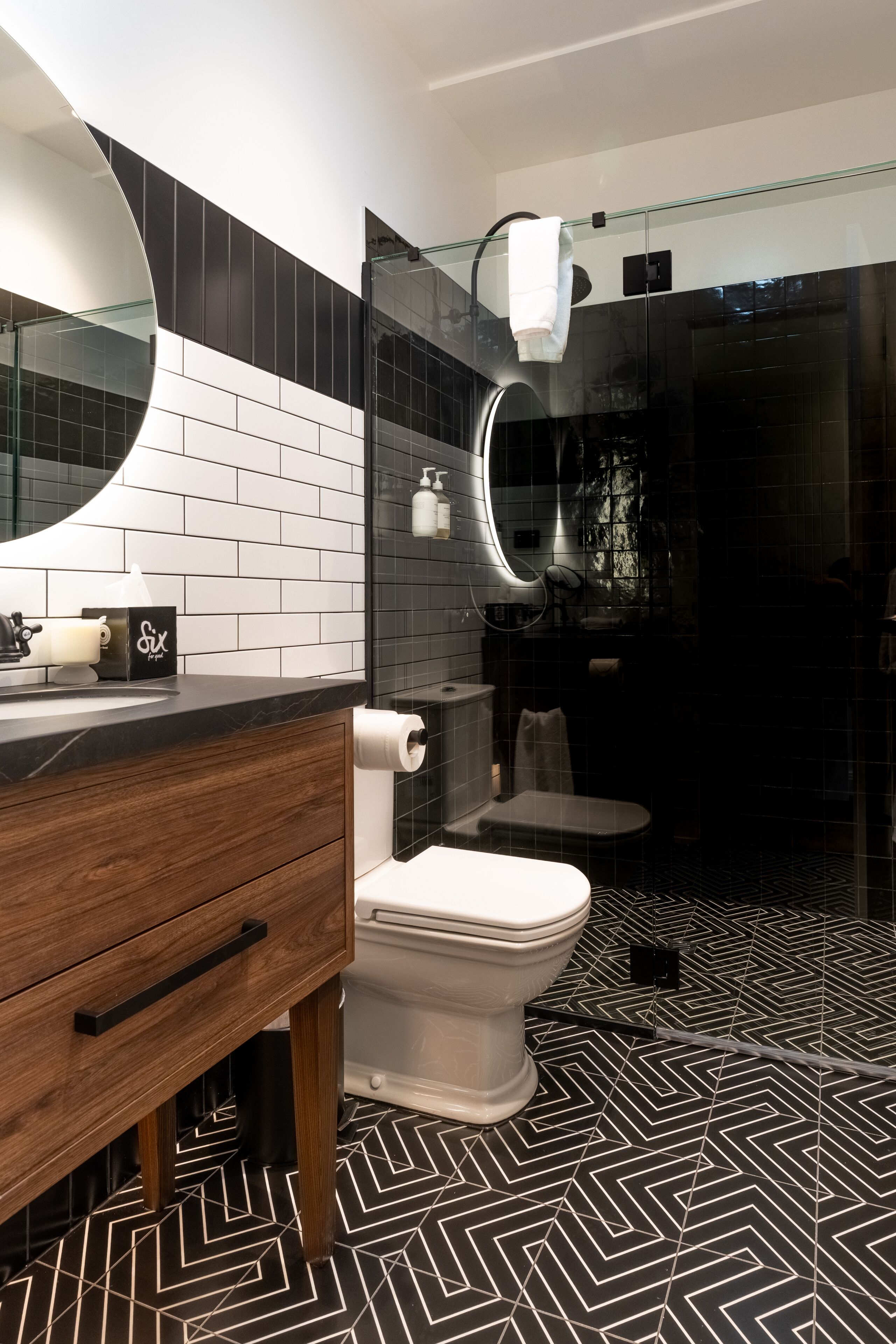 Ein modernes Badezimmer mit schwarz-weißen Fliesen, einem Holzwaschtisch, einer weißen Toilette, einer Duschkabine aus Glas und einem großen Spiegel.