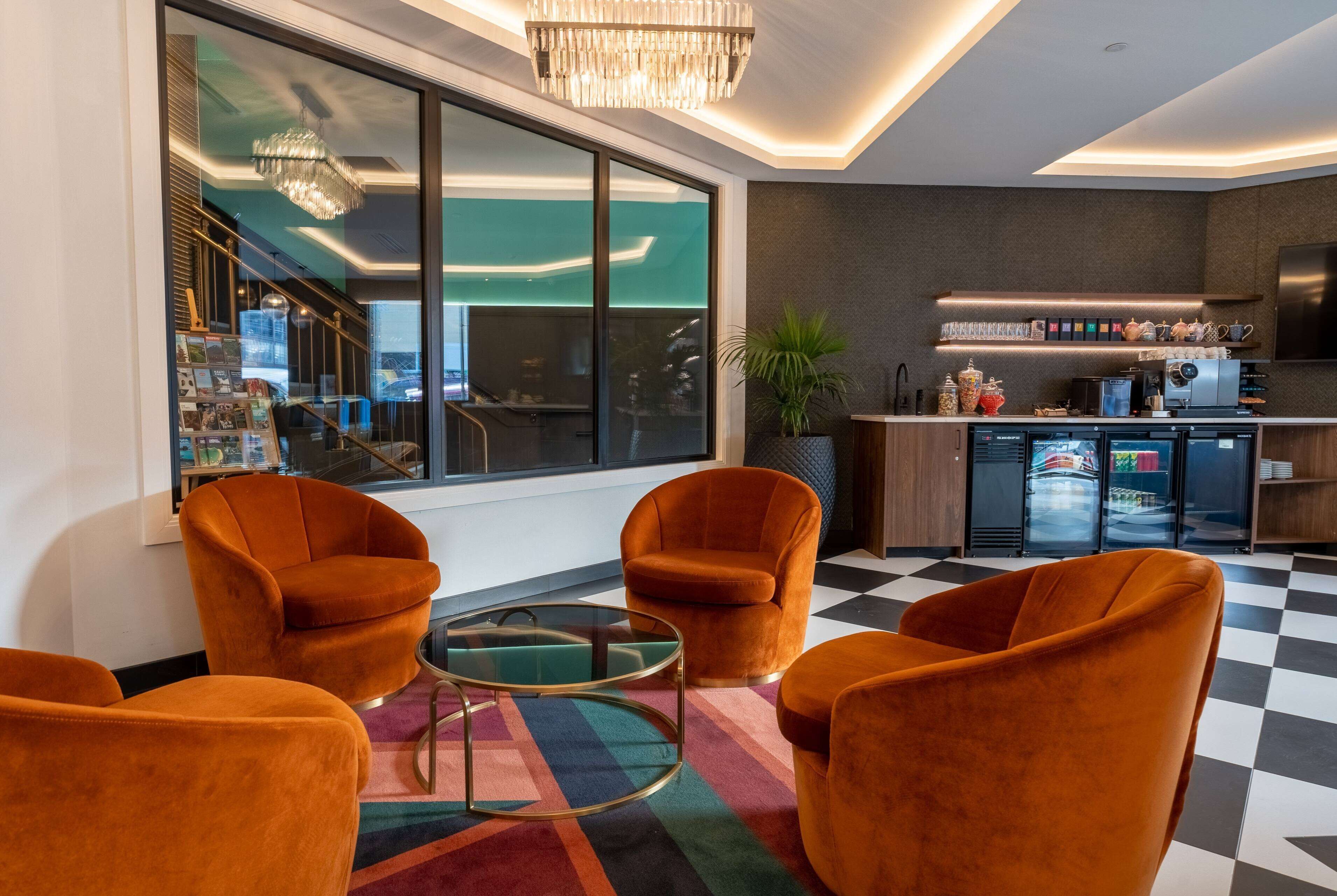 A modern interior with orange velvet armchairs, a glass coffee table, and a checkered floor. A kitchen with a bar area is visible through a glass partition.