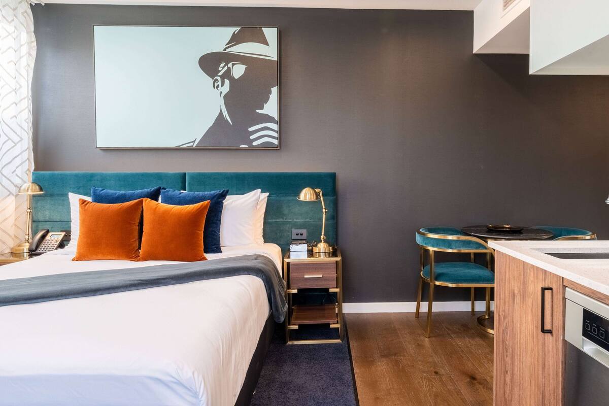 A hotel room with a bed, bedside table, lamp, and a small kitchenette area.
