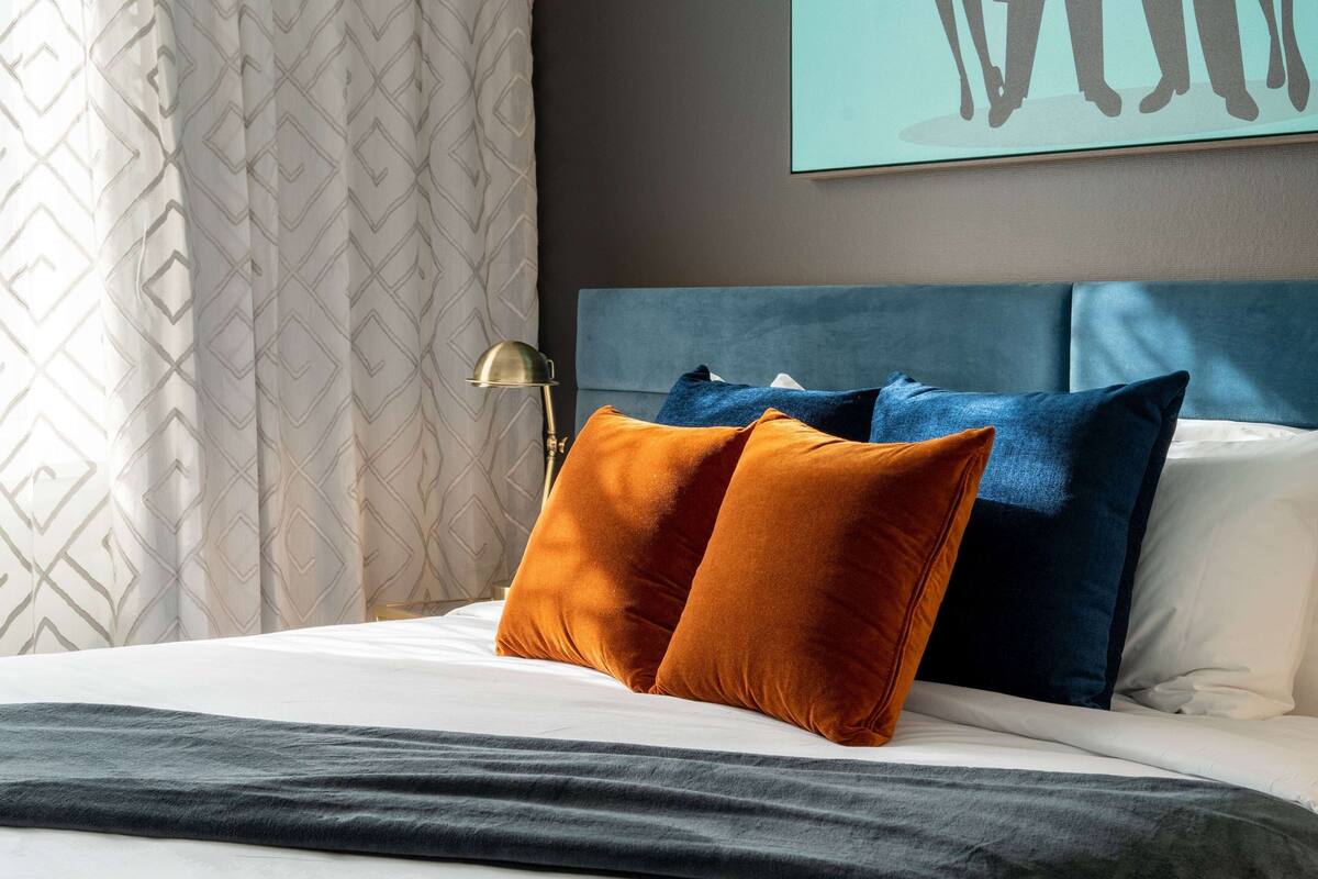 A neatly made bed with two pillows, a bedside lamp, and a geometric-patterned curtain.