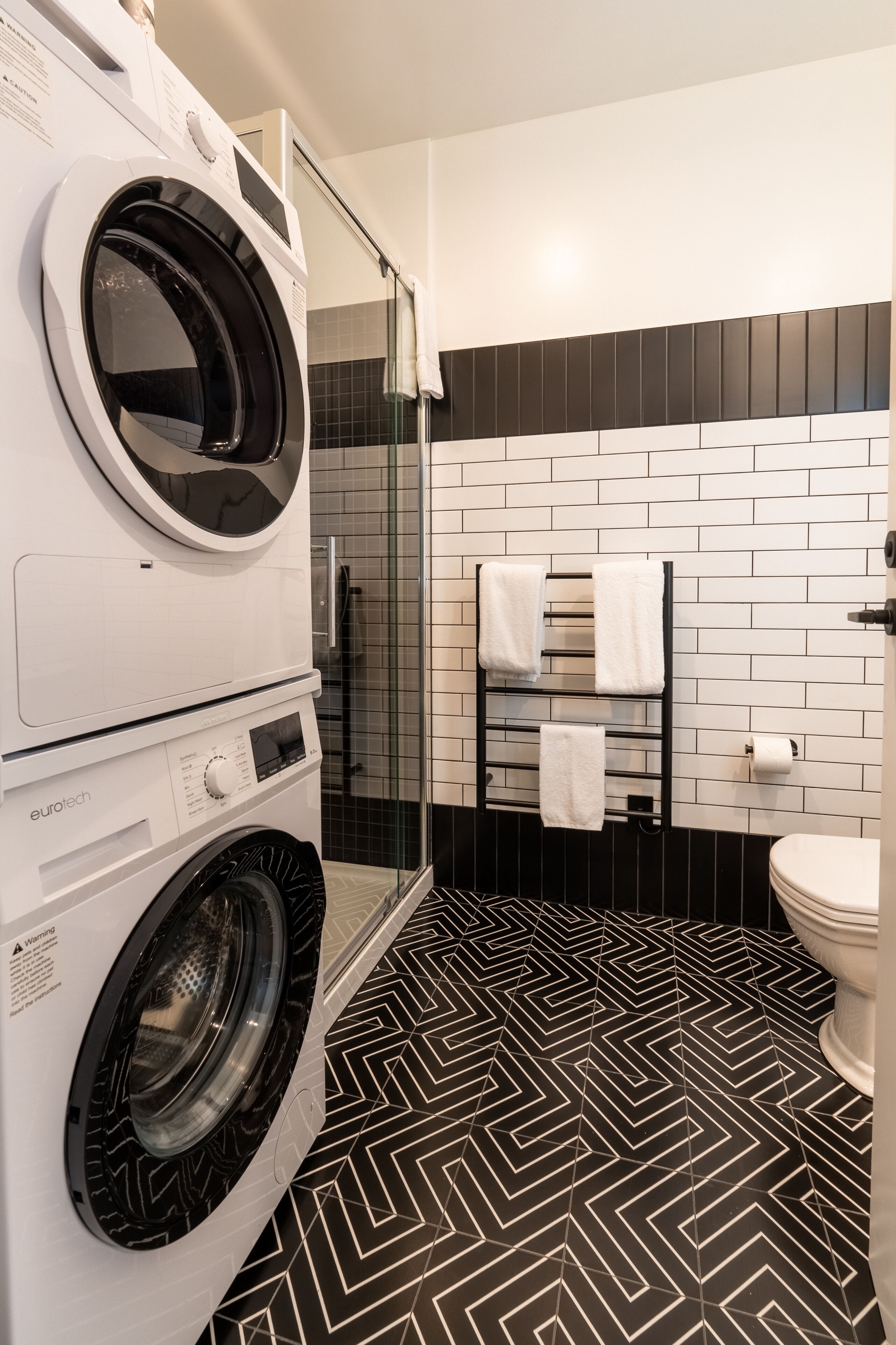 Un bagno moderno con pavimento piastrellato in bianco e nero, lavatrice, box doccia in vetro e un water.