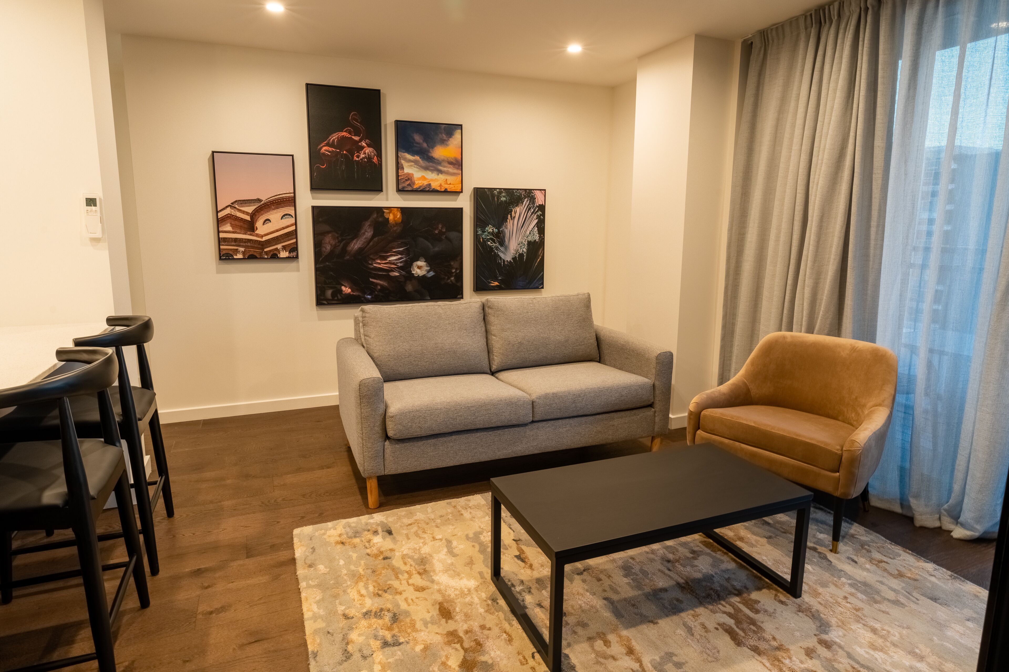 A living room with a grey sofa, a brown armchair, a coffee table, and a dining area with chairs. There are four framed artworks on the wall.