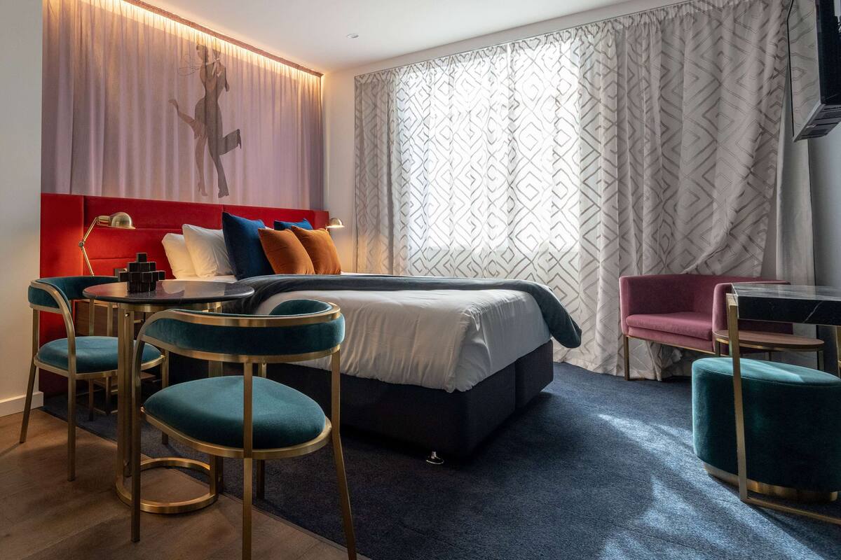 A modern hotel room with a large bed, two bar stools, a small table, a red chair, and a patterned curtain.