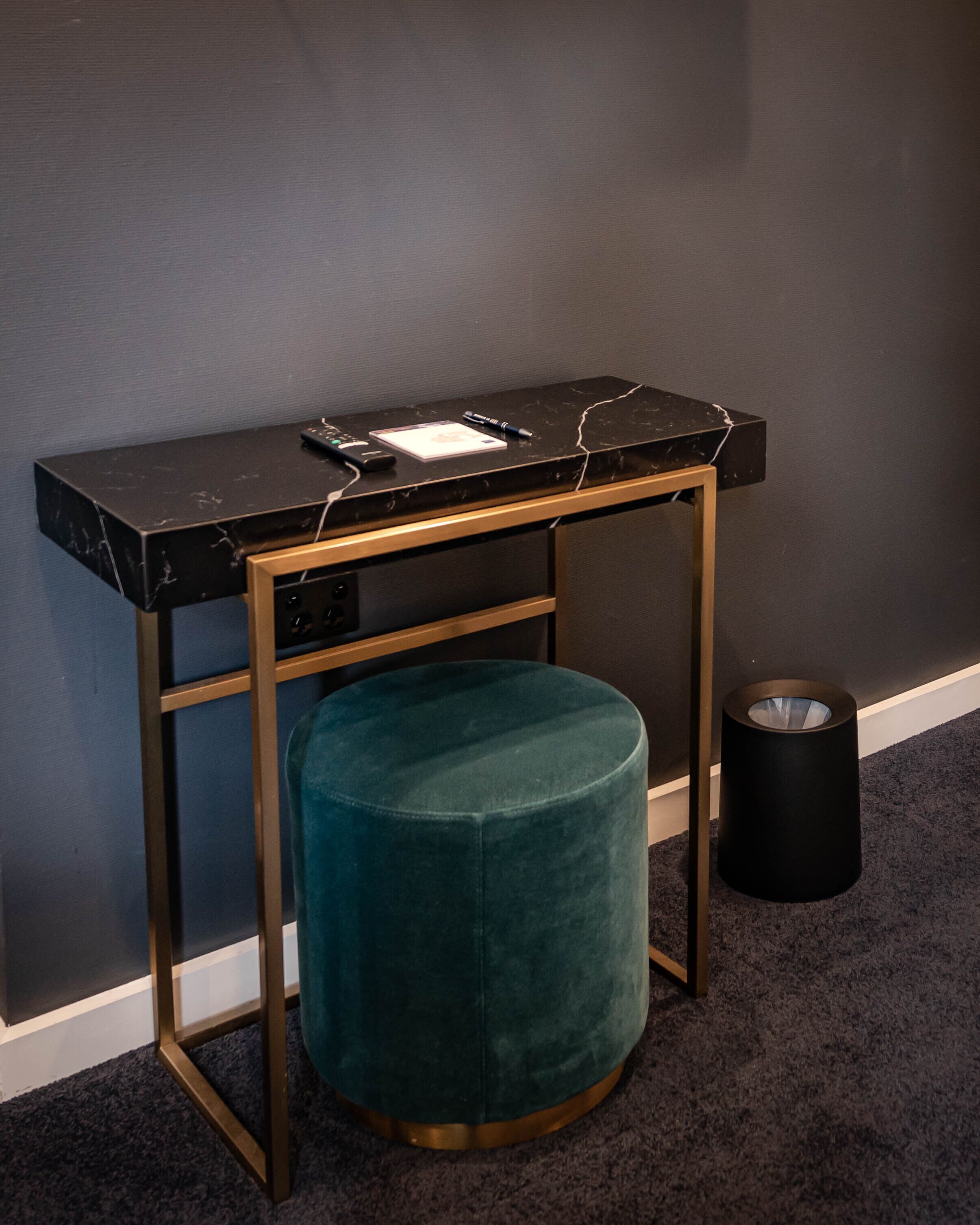 Un bureau en marbre noir avec un cadre doré, un pouf turquoise et une poubelle noire.