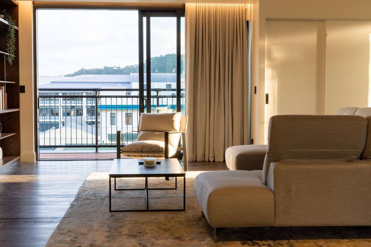 A modern living room with a sofa, armchair, and coffee table. A large window offers a view of a building and greenery.