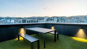 A rooftop terrace with a table and benches, surrounded by a cityscape view.
