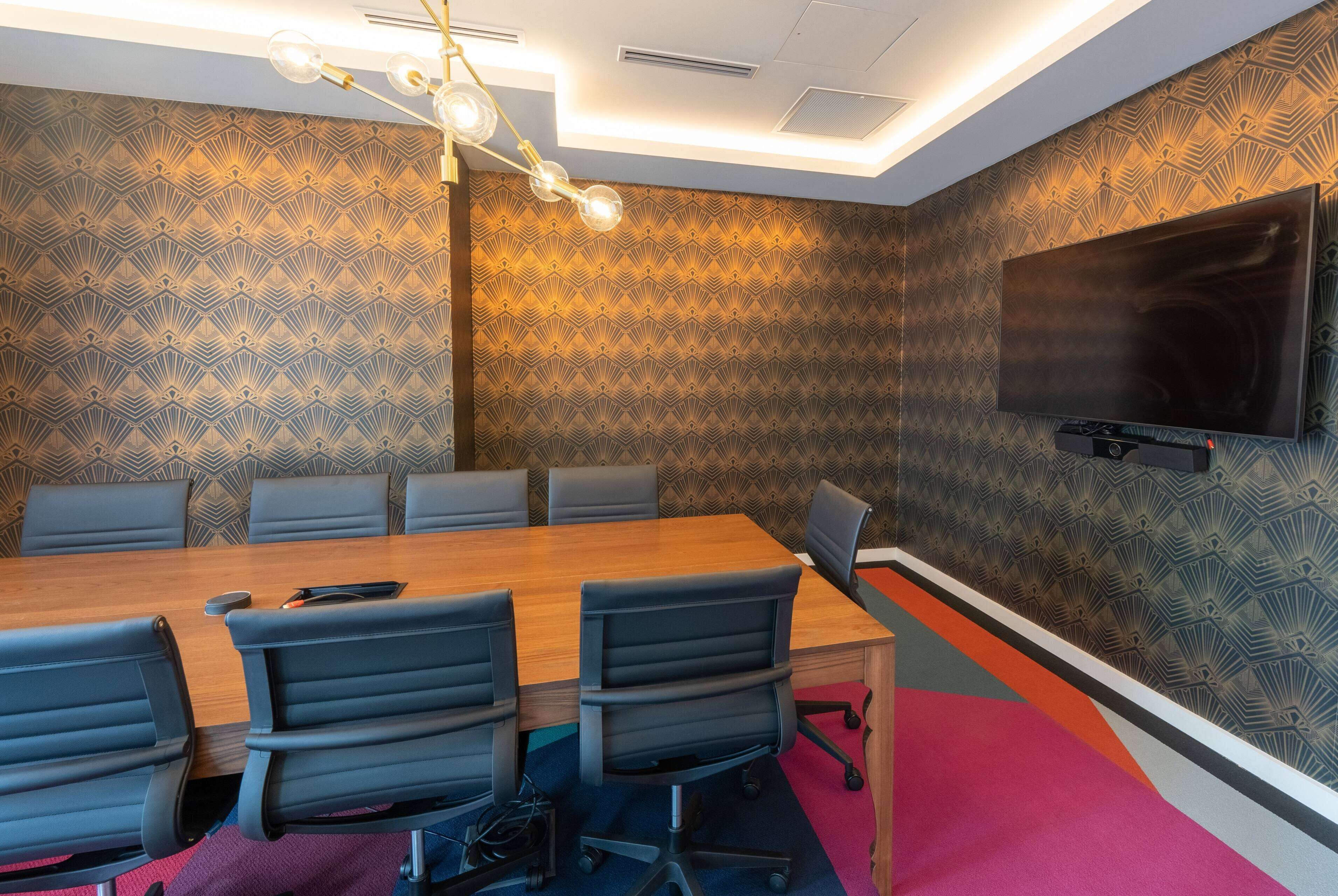 Una sala de conferencias con una gran mesa, seis sillas, un televisor de pantalla plana, papel tapiz estampado y una lámpara en el techo.