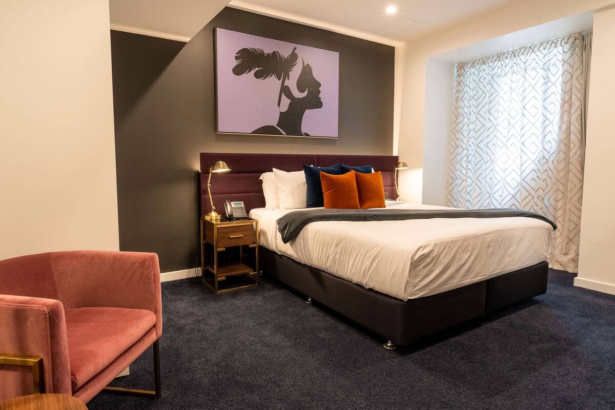 A bedroom with a large bed, a nightstand, a chair, and a wall-mounted artwork.