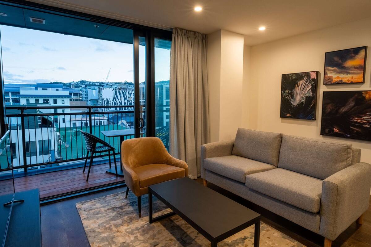 A modern living room with a sofa, armchair, and coffee table, large windows with a view of buildings and a balcony.