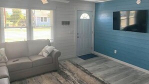 Una habitación con un sofá color crema, un tapete estampado, un ventilador de techo, una puerta y una pared azul con un televisor montado.