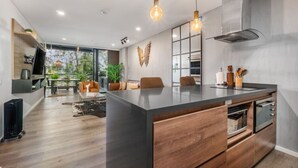 A modern kitchen with a large island, wooden cabinets, and a built-in oven. There is a dining area with a sofa and chairs, and a large window providing natural light.