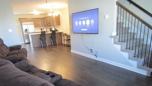 A living room with a large flat-screen TV mounted on the wall, a staircase, and a kitchen area in the background. - Cheerful 2-Bedroom Townhouse with Pool (Clarksville)