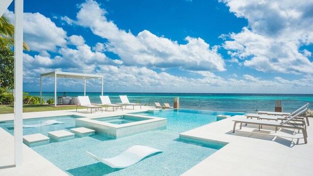 A pool area with clear blue water, white lounge chairs, and a view of the ocean under a partly cloudy sky. - Luxury 6 Bedroom Villa on the Beach in Bodden Town (Breakers)