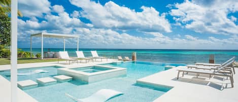 Un área de piscina con agua azul cristalina, sillas de descanso blancas y vistas al océano bajo un cielo parcialmente nublado.