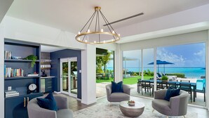 A modern living room with a view of the ocean, a bookshelf, and a dining area with outdoor seating. - Luxury 6 Bedroom Villa on the Beach in Bodden Town (Breakers)